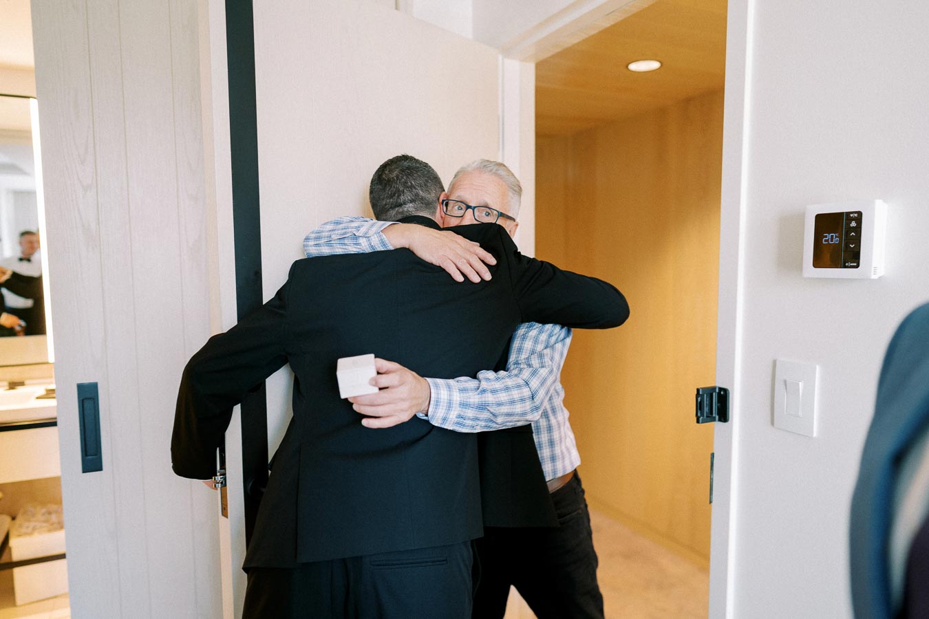 Two men embracing in a heartfelt hug at a doorway, with one holding a small gift box, signifying a special occasion or celebration.