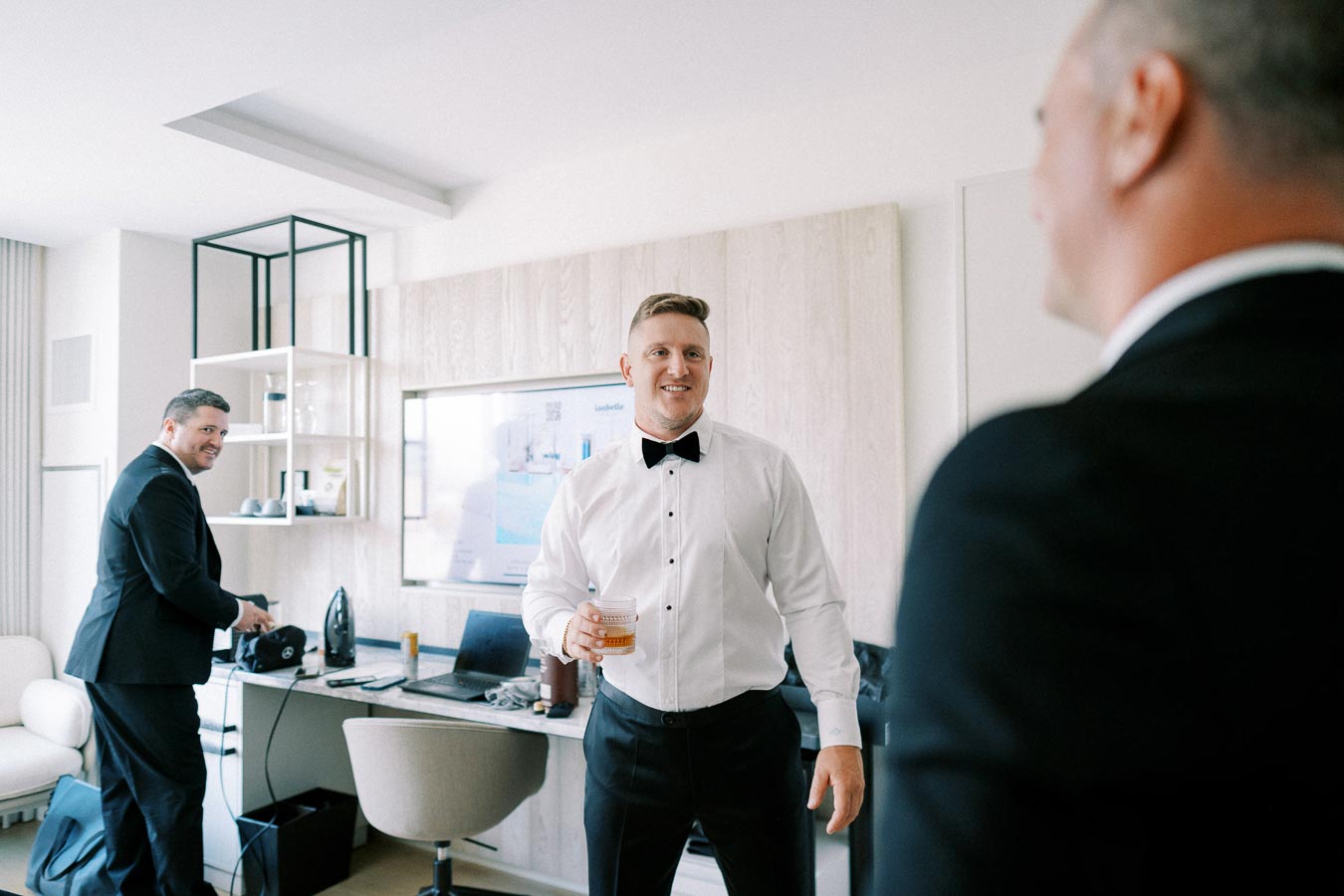 Groom getting ready with friends, wearing formal attire, in a modern hotel room.