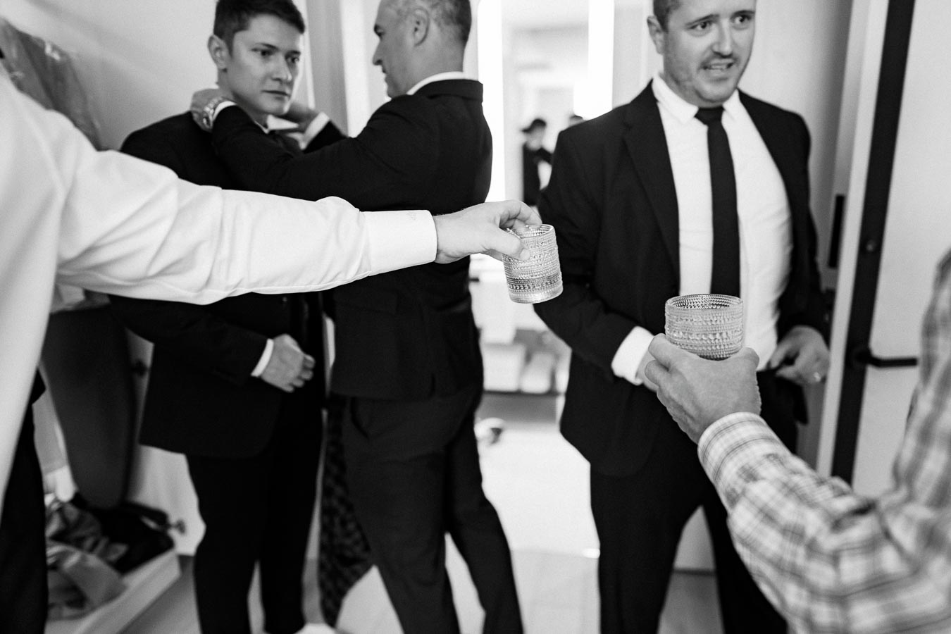 A group of men in suits preparing for a formal event, sharing drinks and adjusting ties, showcasing camaraderie and celebration in a classic black and white setting.