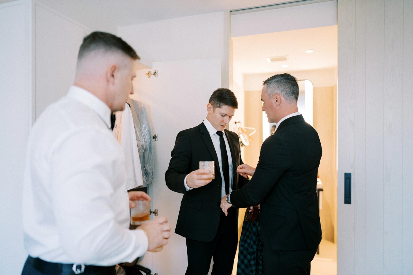 Three men in formal attire preparing for an event, with two adjusting suits and one holding a drink, in a well-lit room.