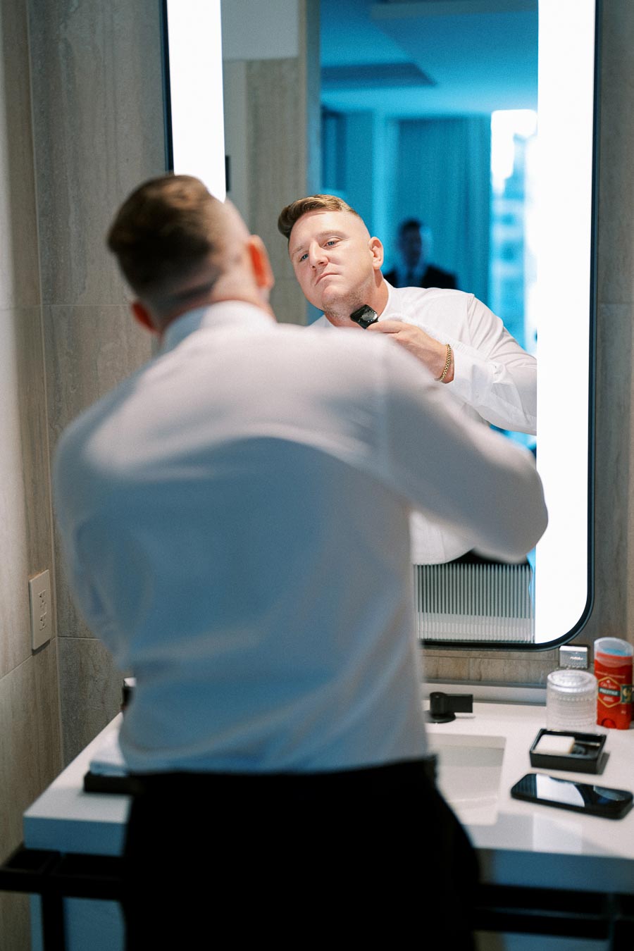 A man in a white shirt grooming himself in front of a bathroom mirror, focusing on shaving with an electric razor.