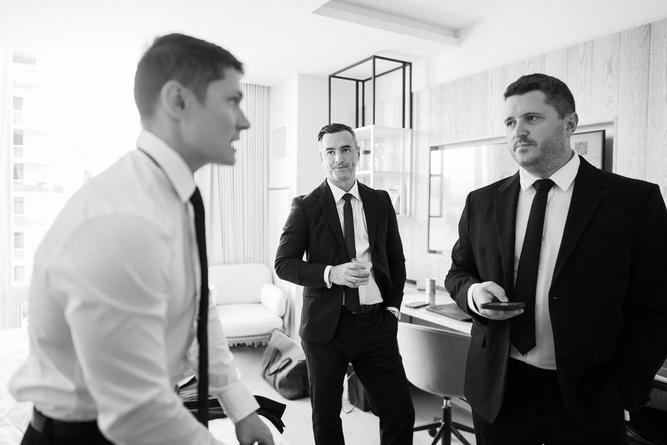 Black and white image of three men in suits conversing in a modern office setting, one holding a drink.