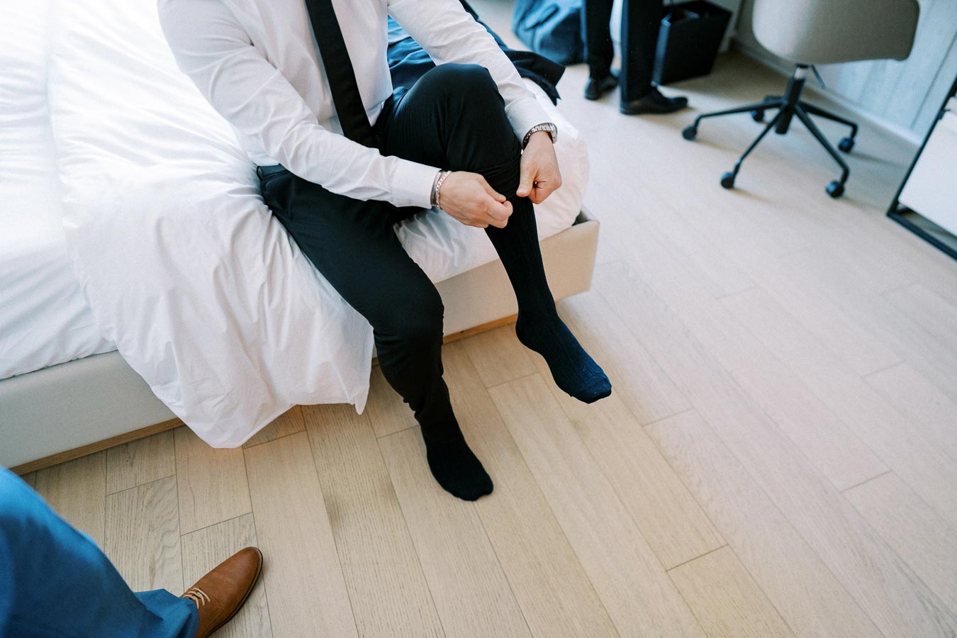 A man in formal attire sitting on the edge of a neatly made bed, adjusting his black socks in a well-lit, modern bedroom with light wood flooring.