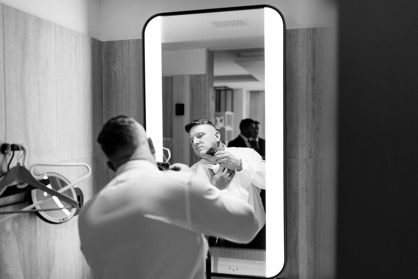 A man adjusting his tie in front of an illuminated mirror in a bathroom, preparing for a formal event, with wooden paneling and hangers visible in the background.