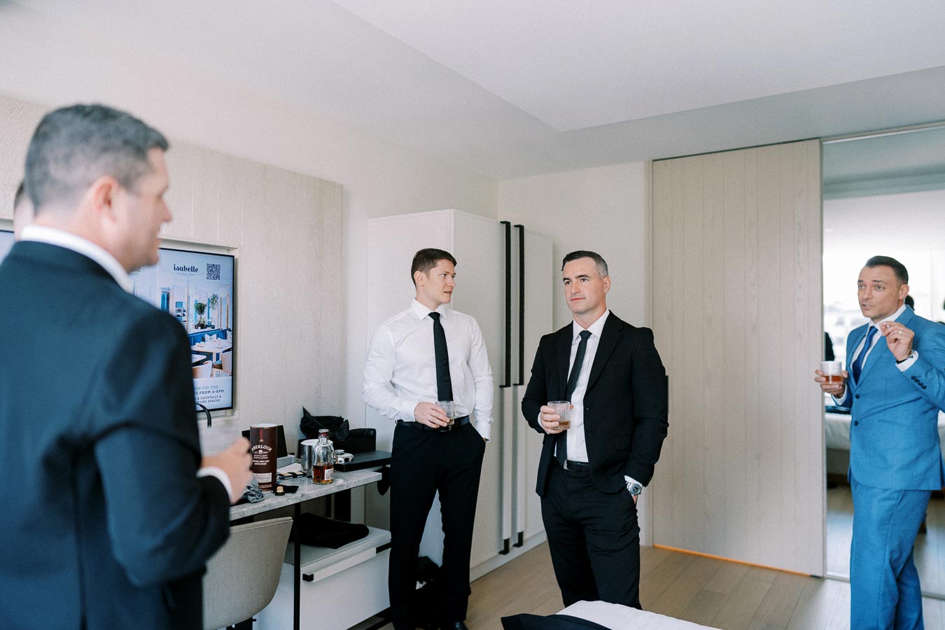 Four men in suits standing and talking in a modern hotel room, holding drinks, with a focus on professional attire and casual conversation atmosphere.