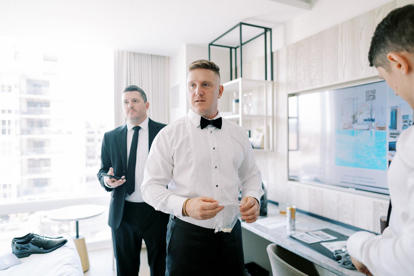 Sure! Here's an SEO-optimized alt text for the image:Formal attire preparation with groom and groomsmen in a modern hotel room, featuring polished dress shoes and stylish suits, creating an elegant wedding day ambiance.