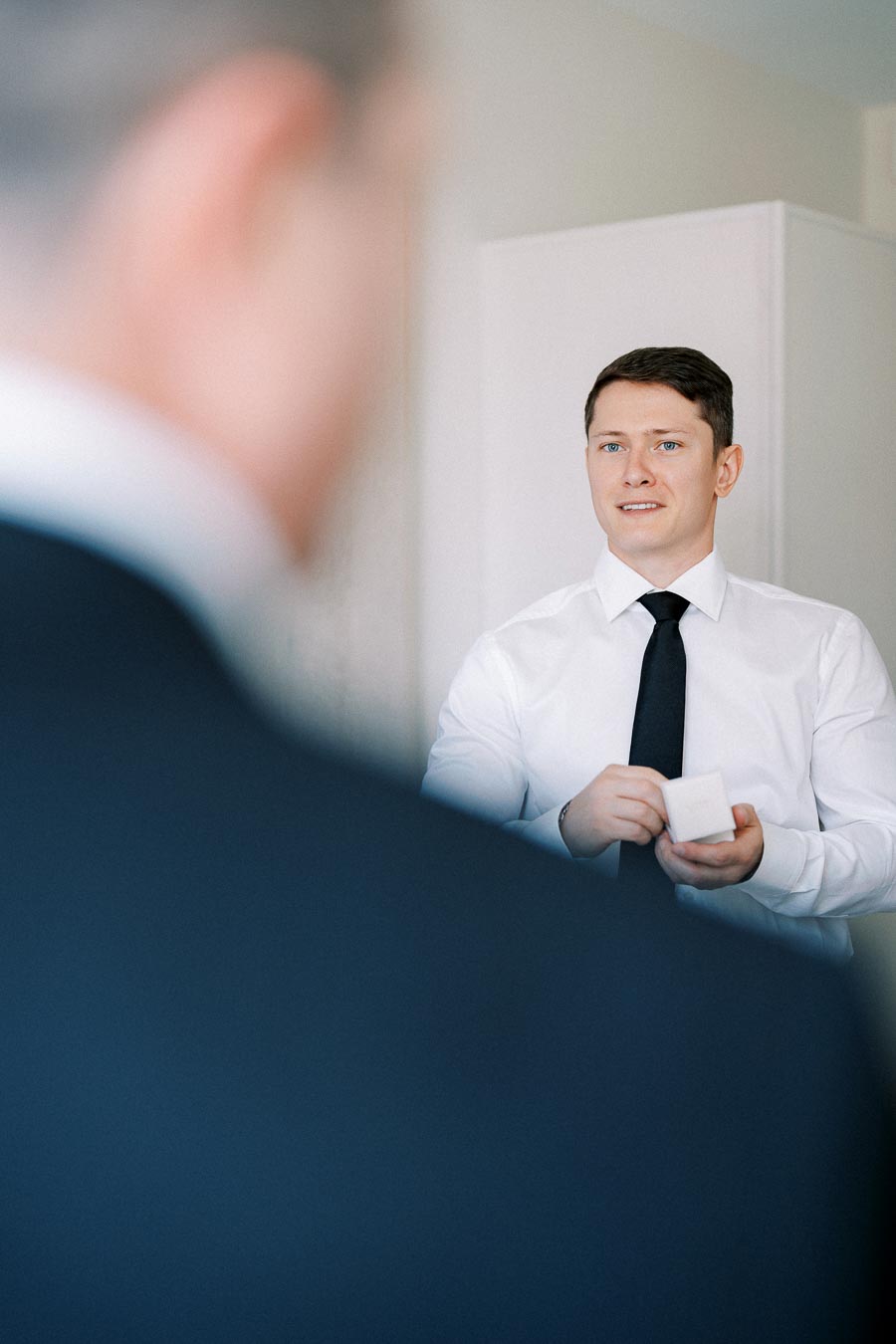 A man in a white shirt and black tie holding a small white box, preparing for a formal occasion, with a blurred figure in the foreground.