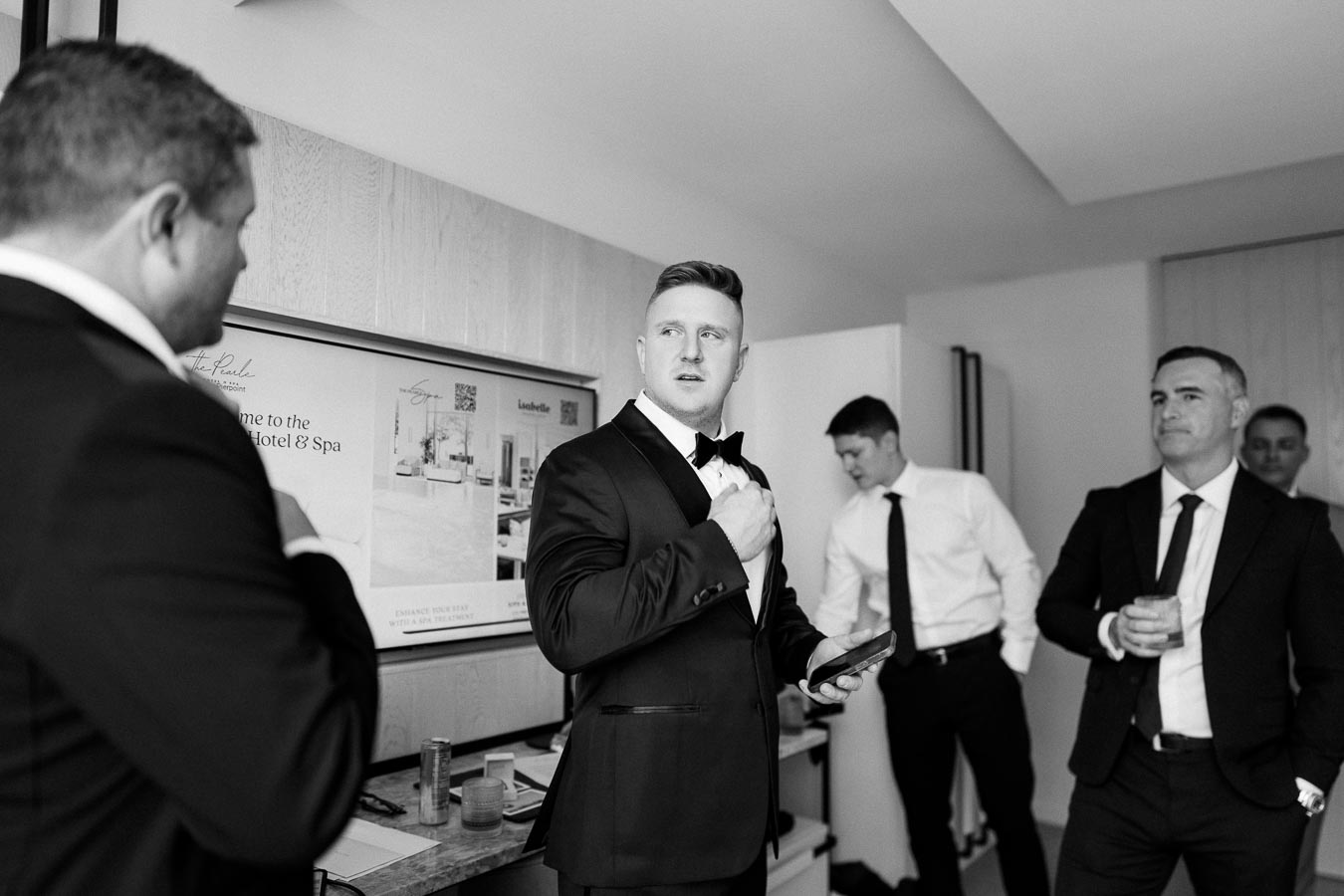 Black and white photo of groom adjusting tie in hotel room with groomsmen preparing for wedding.