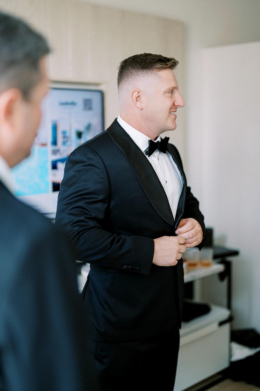 A groom in a black tuxedo preparing for his wedding, buttoning his jacket with a smile.