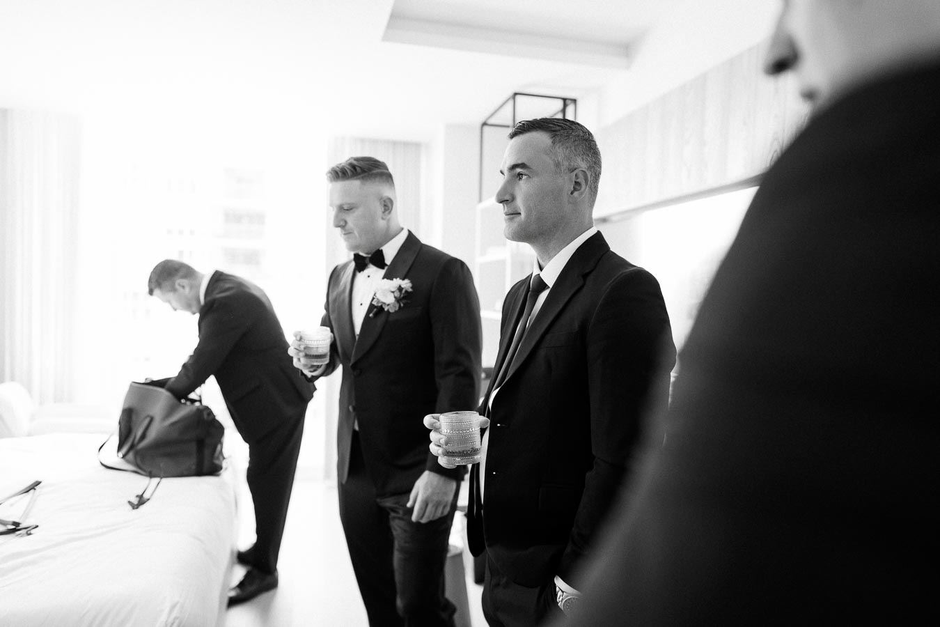 Black and white image of groomsmen in suits preparing for a wedding, one holding a drink, in a bright hotel room setting.