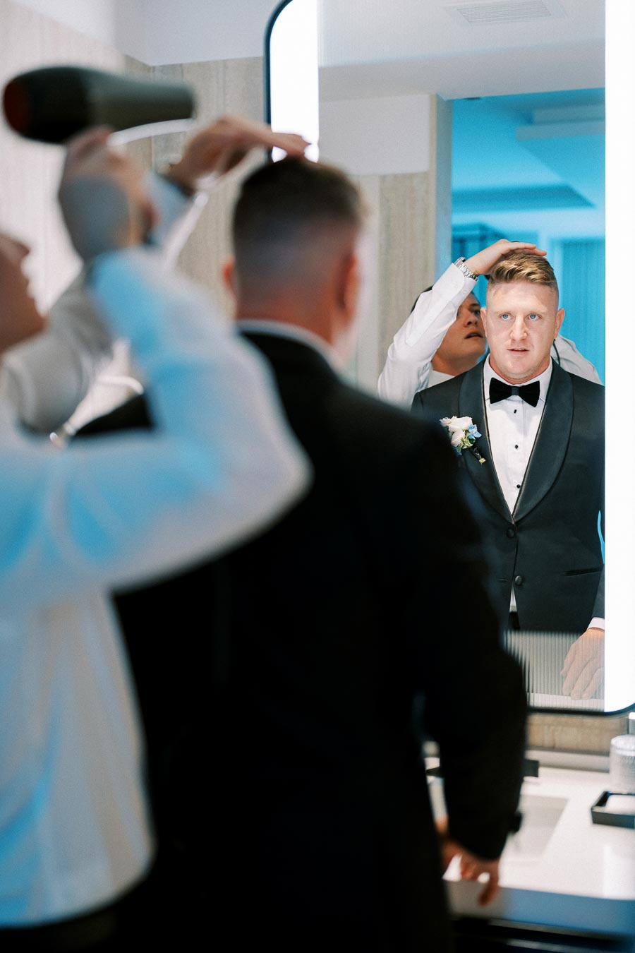 Groom getting ready for wedding, man in tuxedo looks in mirror while stylist adjusts his hair in bathroom.