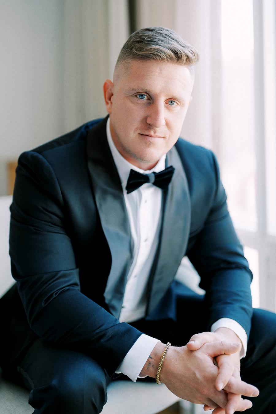 Man in elegant black tuxedo with bowtie sitting indoors, hands clasped, looking confidently at the camera.
