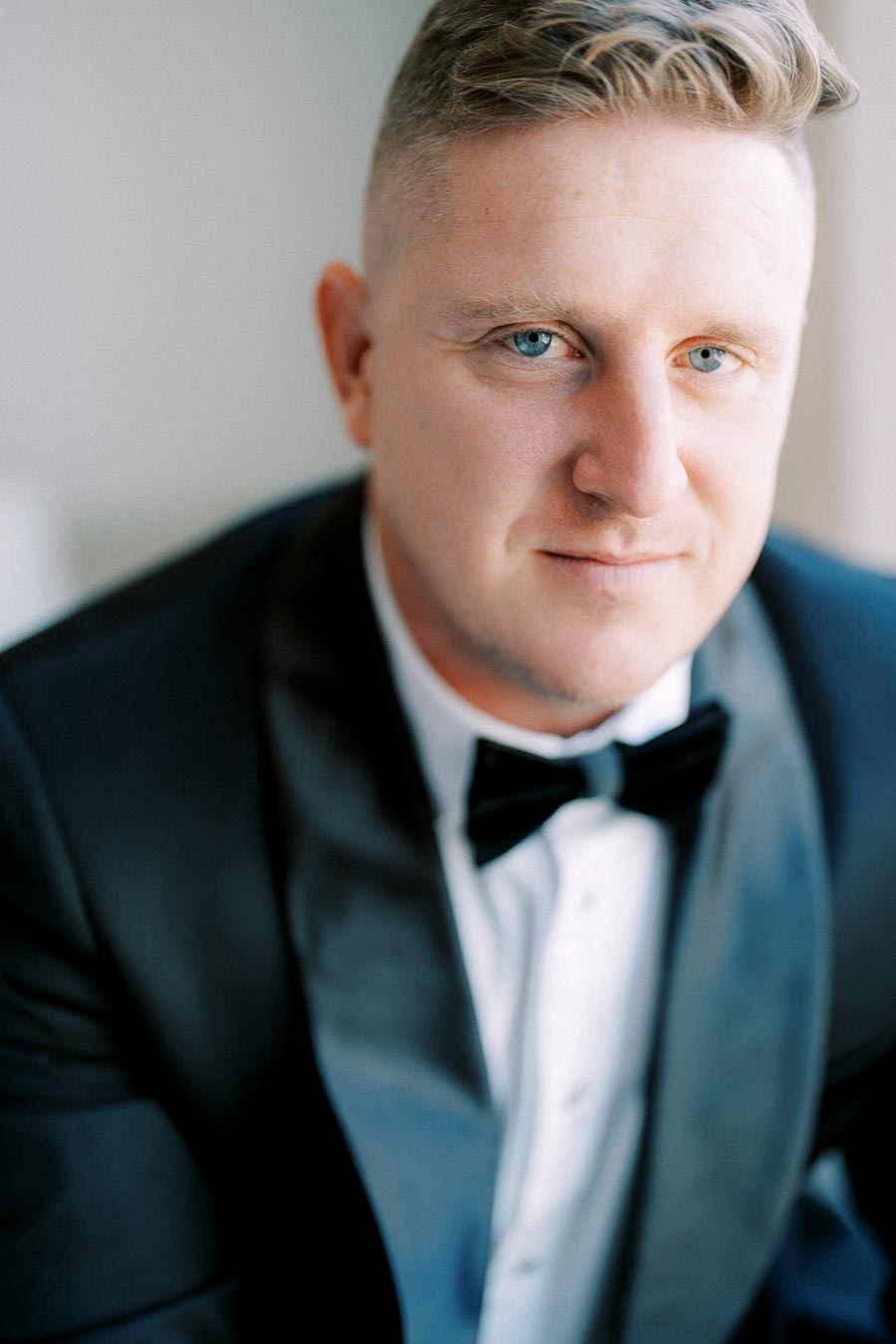 A man wearing a classic black tuxedo with a bow tie, looking directly at the camera with a confident expression, against a softly lit background.