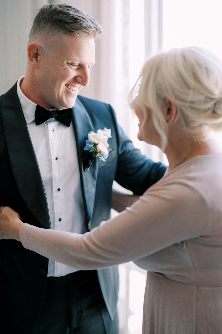 A groom in a tuxedo smiles lovingly at an older woman, possibly his mother, as she adjusts his boutonniere before a wedding ceremony.