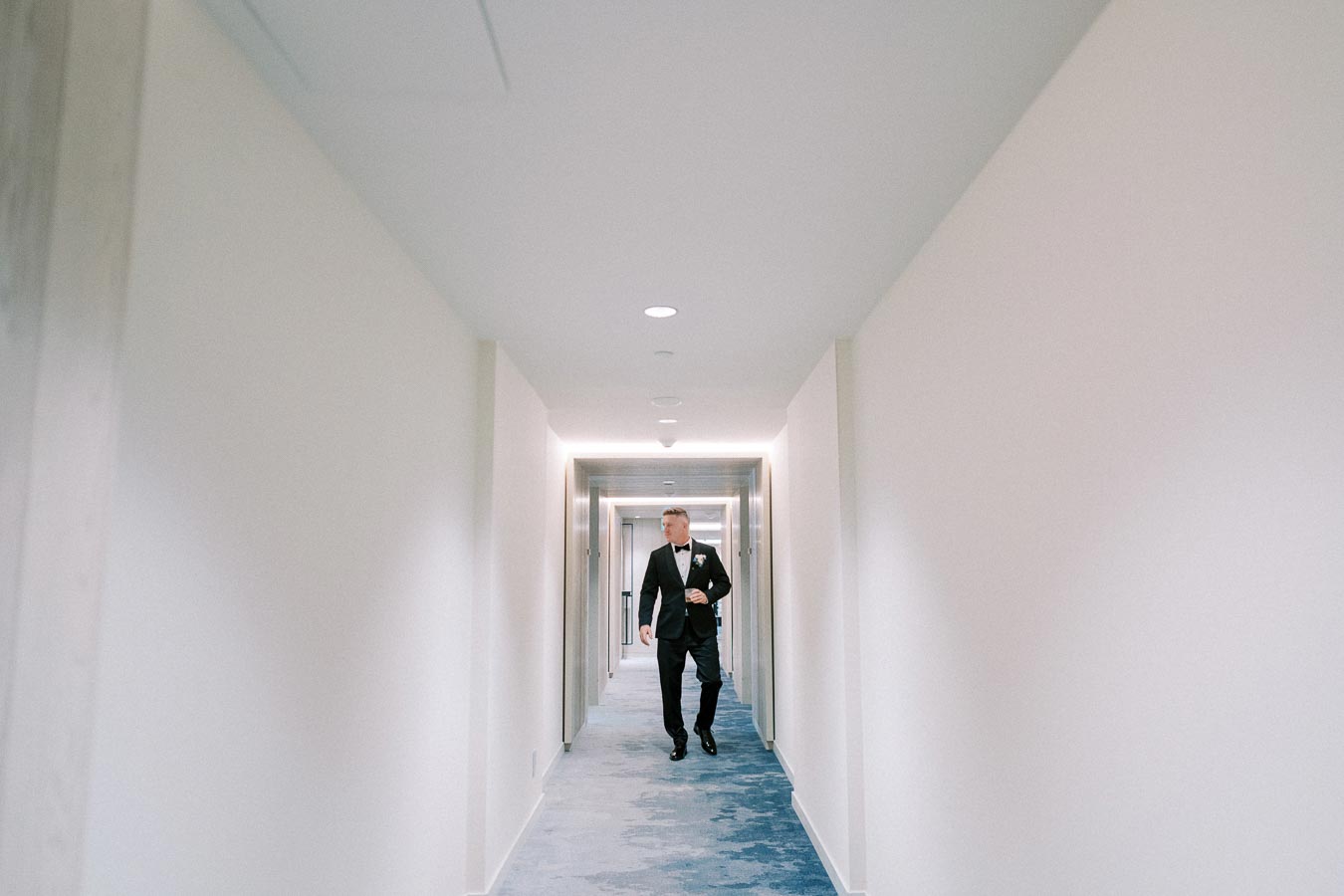 A person in a tuxedo walking down a brightly lit hotel hallway with blue and white carpeting.