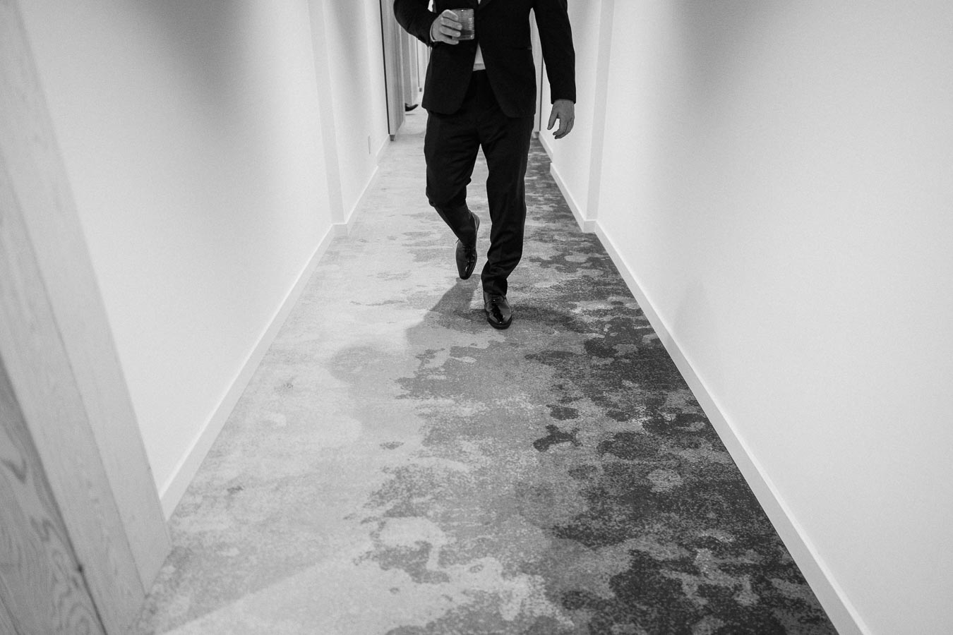Man in a suit walking down a hallway with modern patterned carpet, holding a drink.