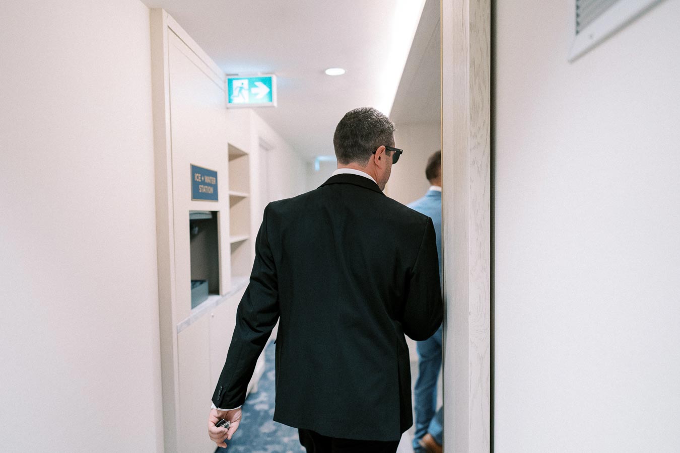 A person in a black suit walking down a corridor with an ice and water station, holding sunglasses in hand, near an emergency exit sign.