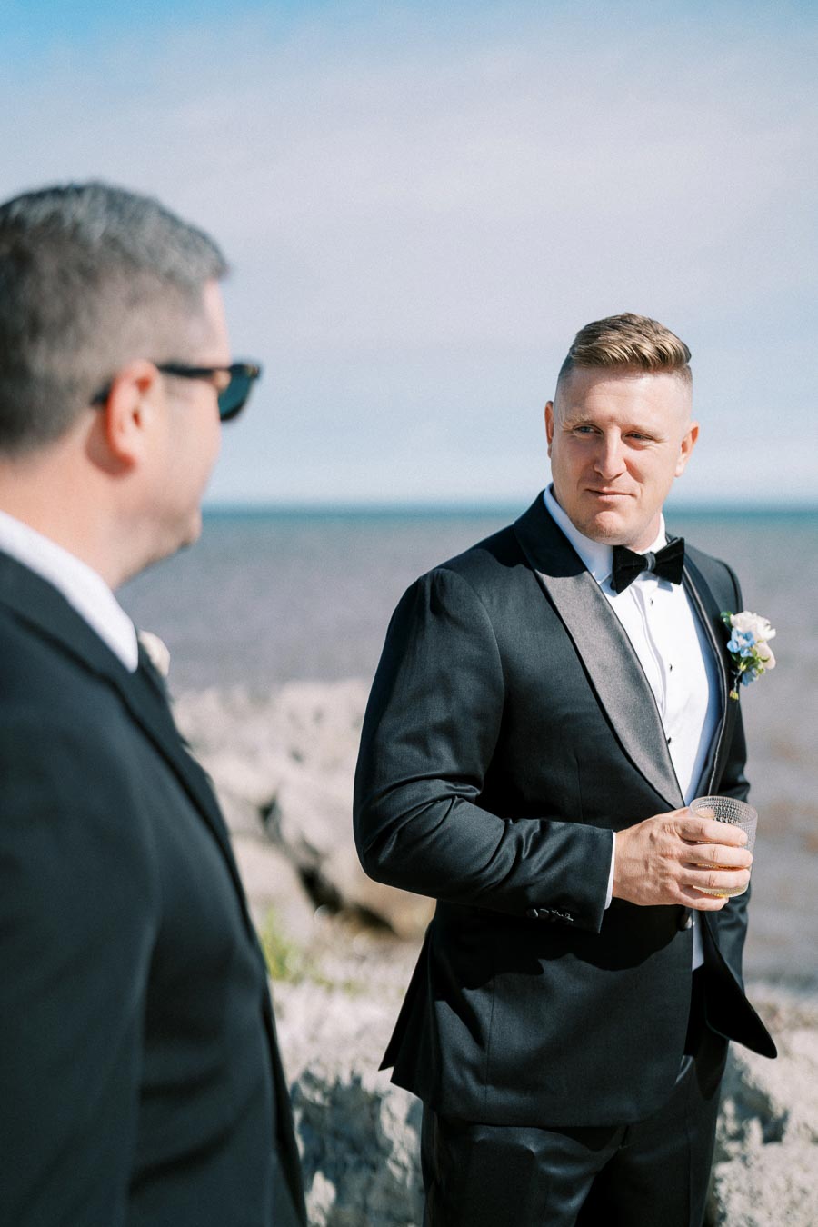 Groom in a black tuxedo holding a drink, standing outdoors near a rocky shoreline, conversing with a man in sunglasses.