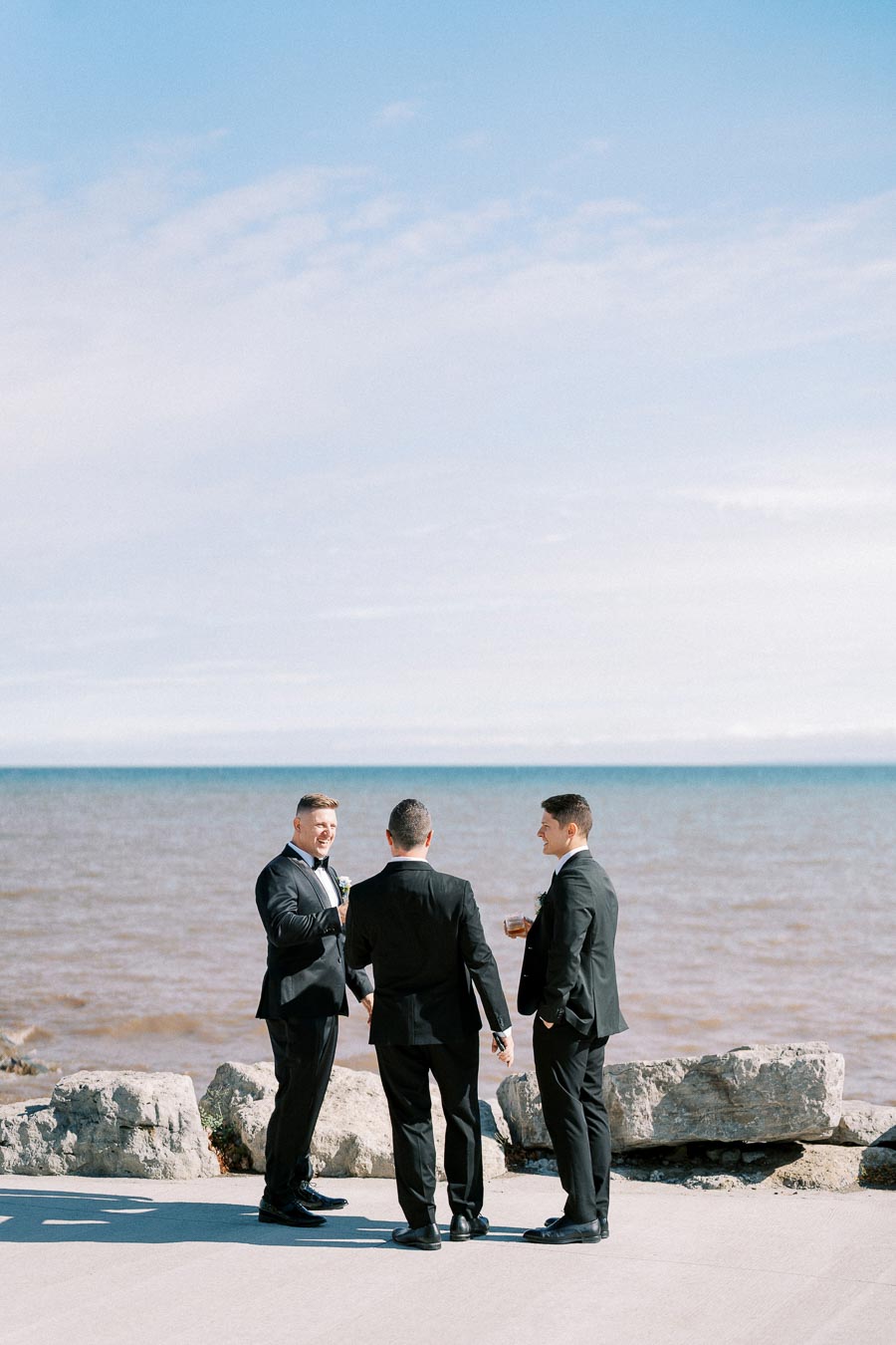 Three men in suits conversing by the ocean on a sunny day, with a clear blue sky and rocky shoreline in the background.