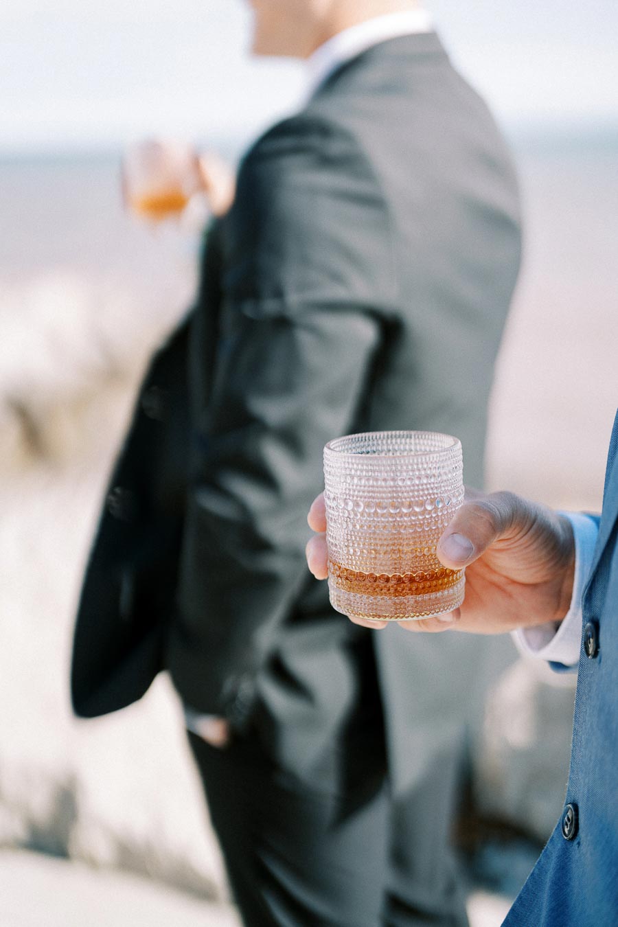 A person in a suit holding a textured glass of whiskey outdoors, with another individual in the background, partially blurred, also in formal attire.