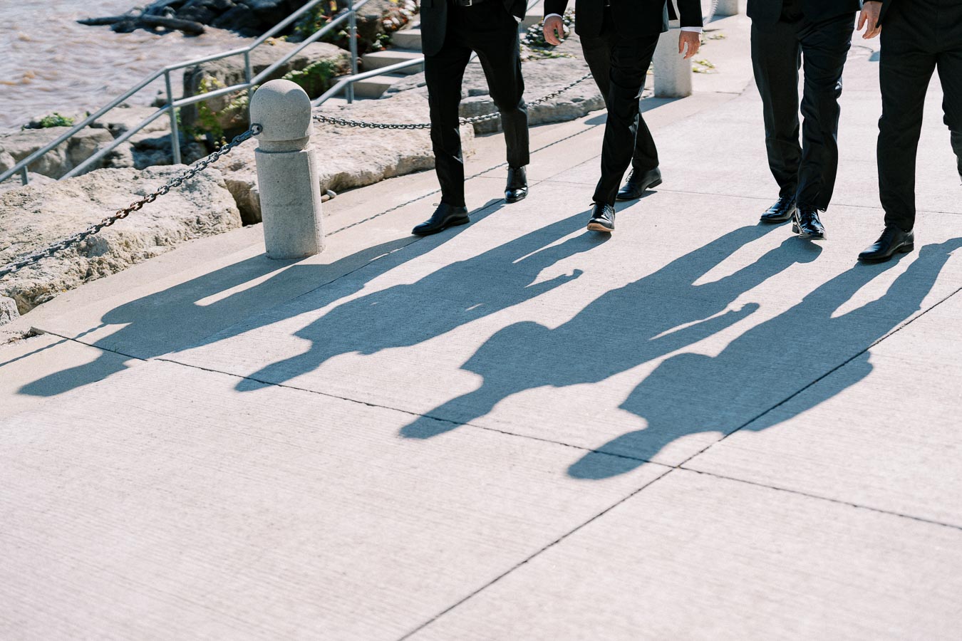 A group of men in suits walking on a sunny waterfront path, casting distinct shadows on the pavement.