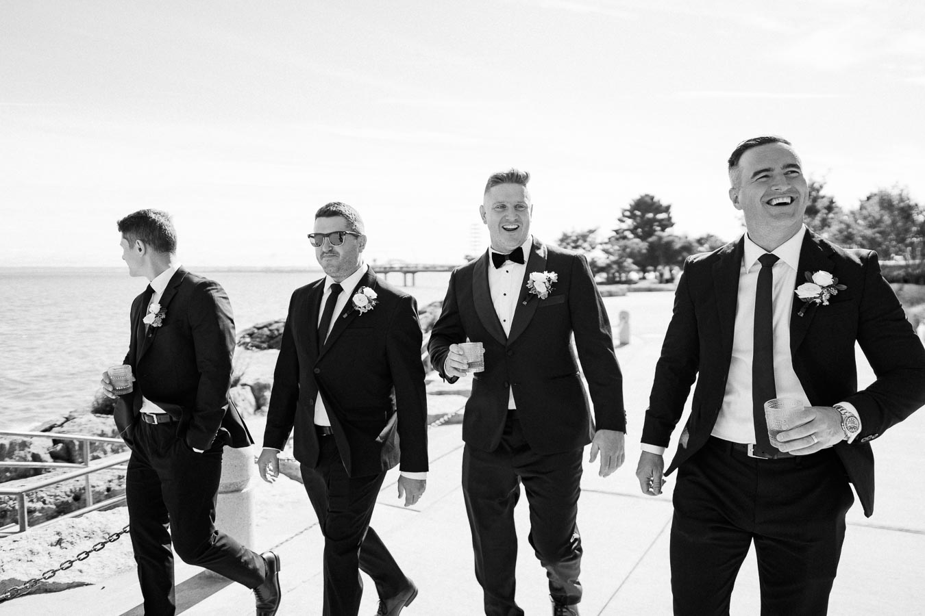Black and white photo of four groomsmen in suits with floral boutonnieres, enjoying drinks while walking along a waterfront on a sunny day.