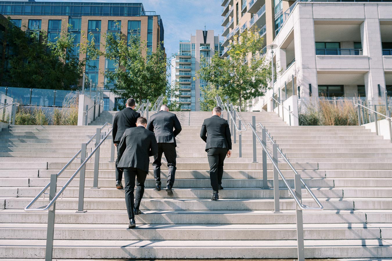 Four men in black suits walking up concrete stairs in an urban setting with modern buildings and trees in the background.