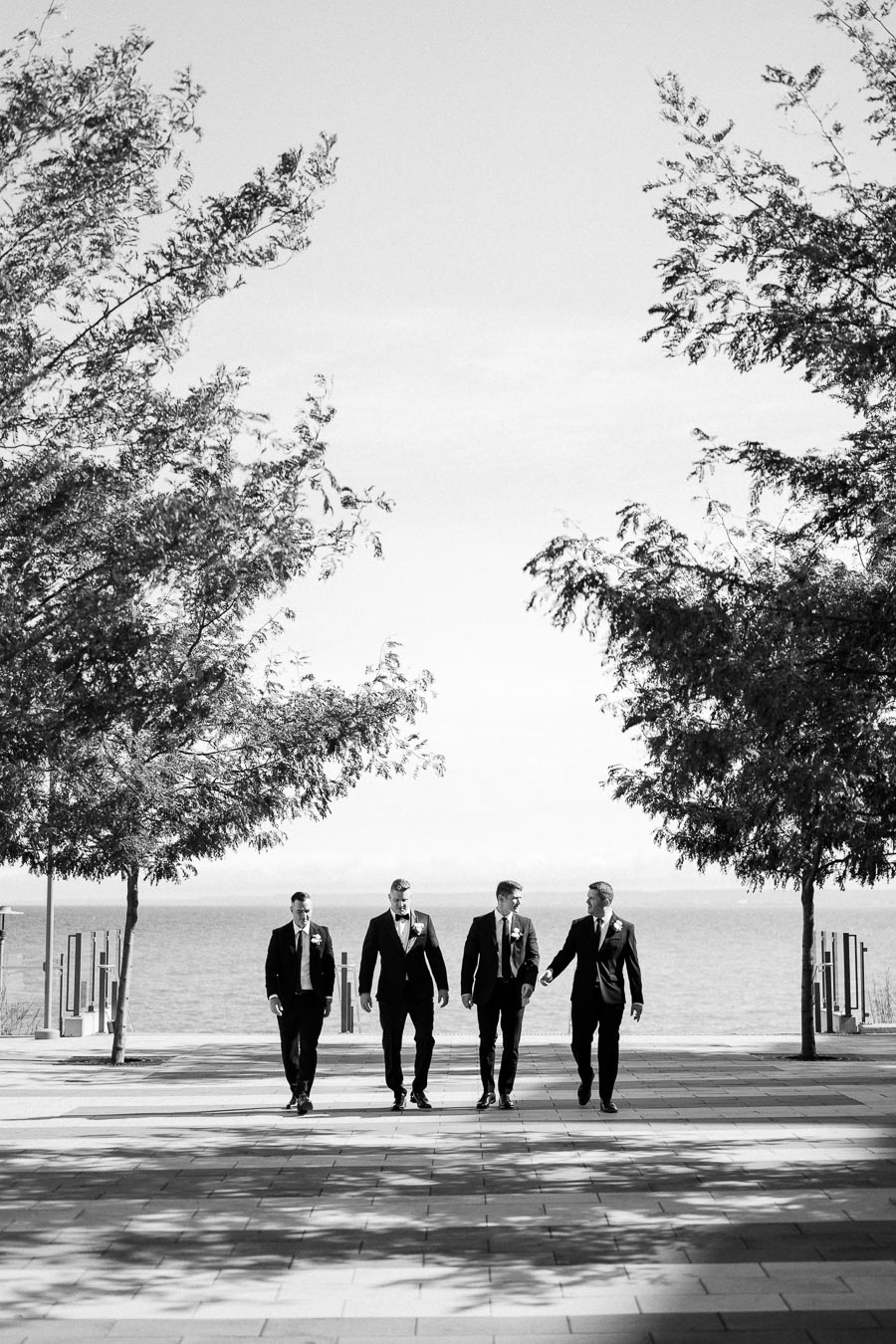 Black and white image of four men in formal suits walking down a tree-lined path near a waterfront.