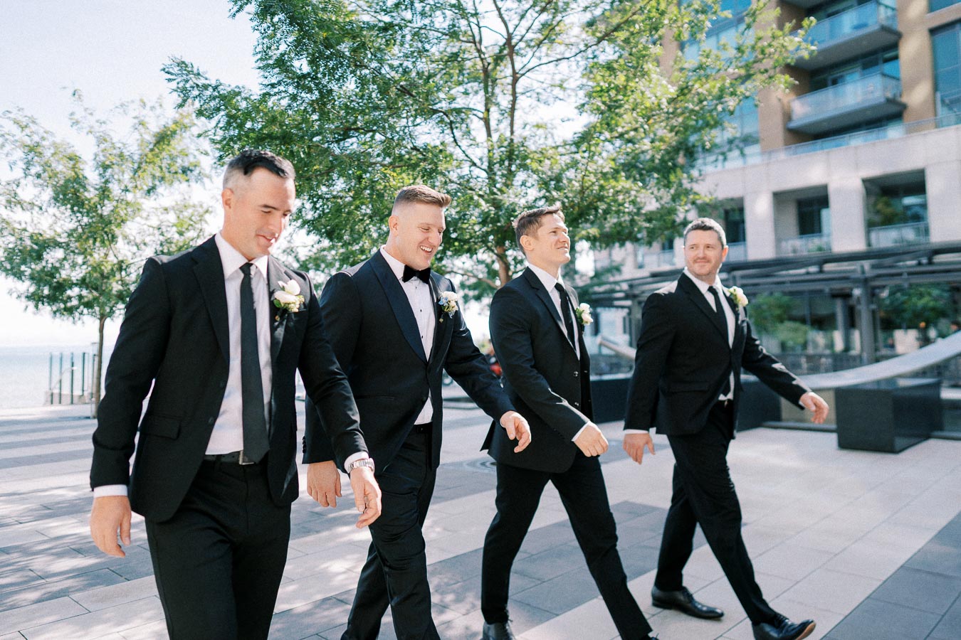 Groomsmen in black suits walking happily outdoors, wearing boutonnières, with trees and a modern building in the background on a sunny day.