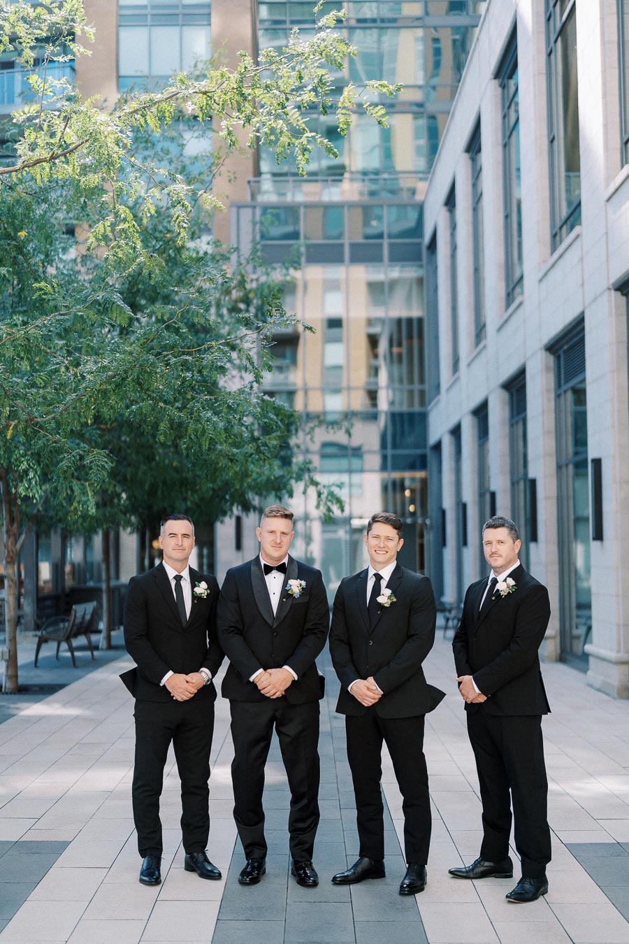 Four groomsmen in black suits stand together for a formal wedding photograph in an urban setting with modern architecture and greenery in the background.