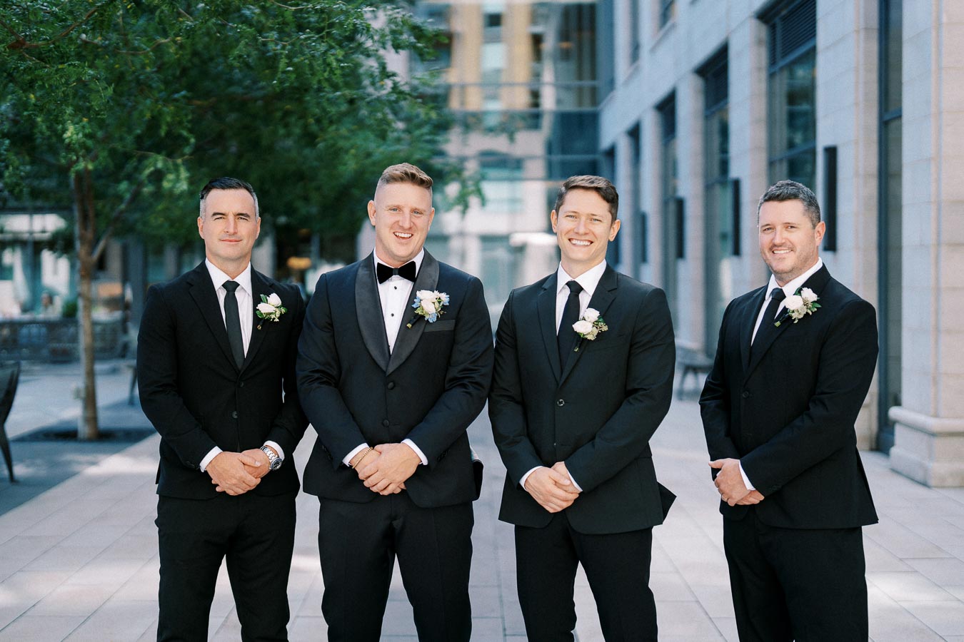 Four groomsmen in black suits with boutonnières posing outdoors, in front of a city building, smiling for the camera.