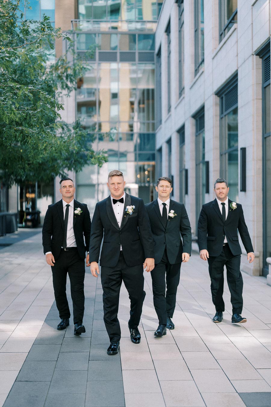 Four men in suits with boutonnieres walking confidently in an urban setting, likely part of a wedding party with the groom at the forefront, modern city architecture in the background.