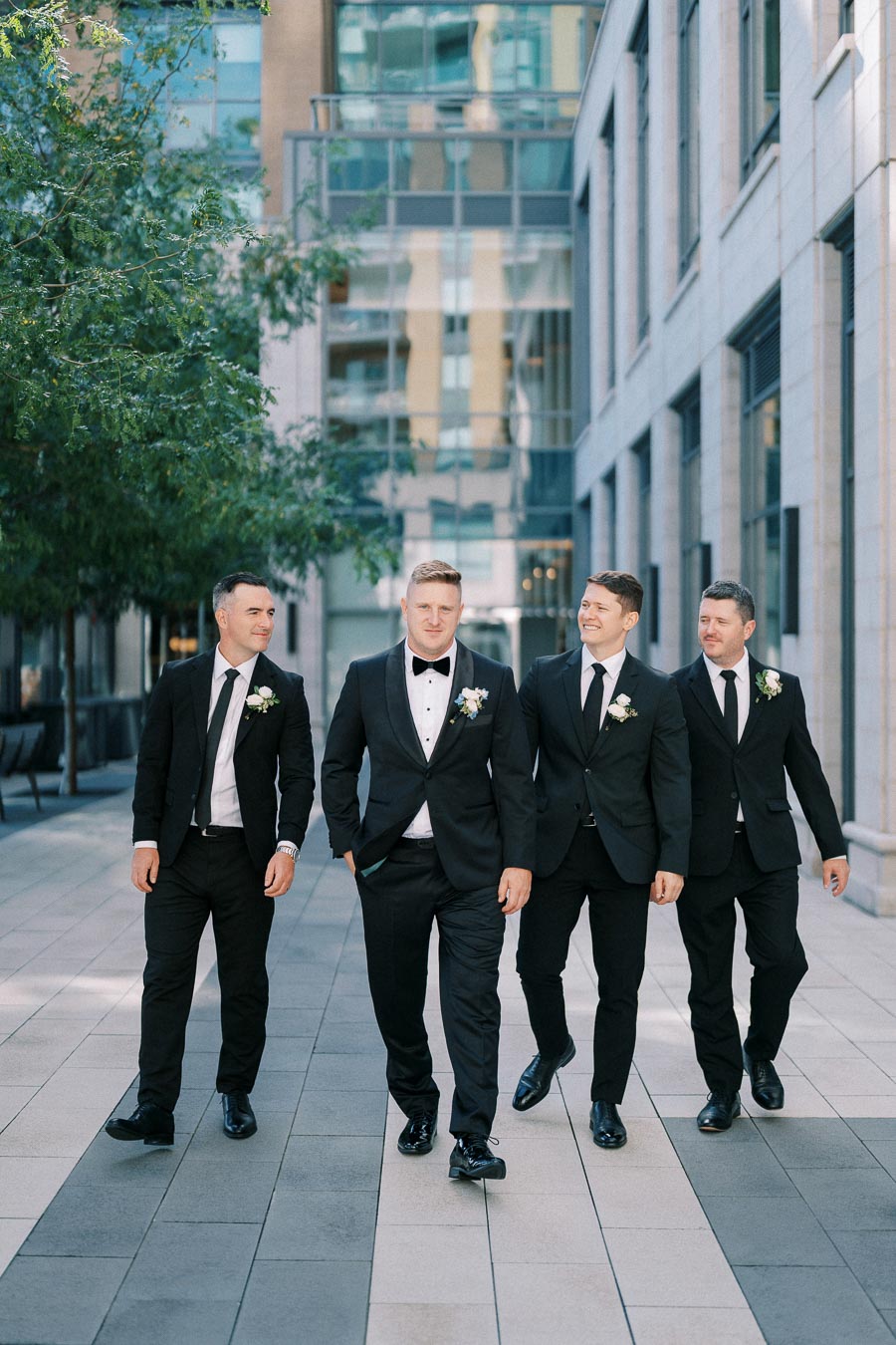 Groom and groomsmen walking in a city setting, dressed in formal black suits and matching ties, with boutonnières, showcasing a modern wedding fashion theme.