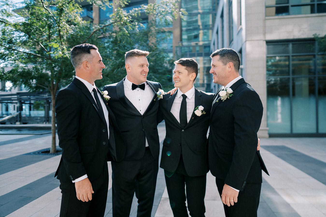 Four groomsmen in formal suits smile and embrace each other outdoors near modern buildings.