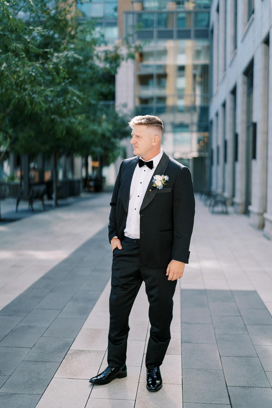Man in a black tuxedo with a bow tie standing on an urban street, surrounded by modern buildings and greenery.