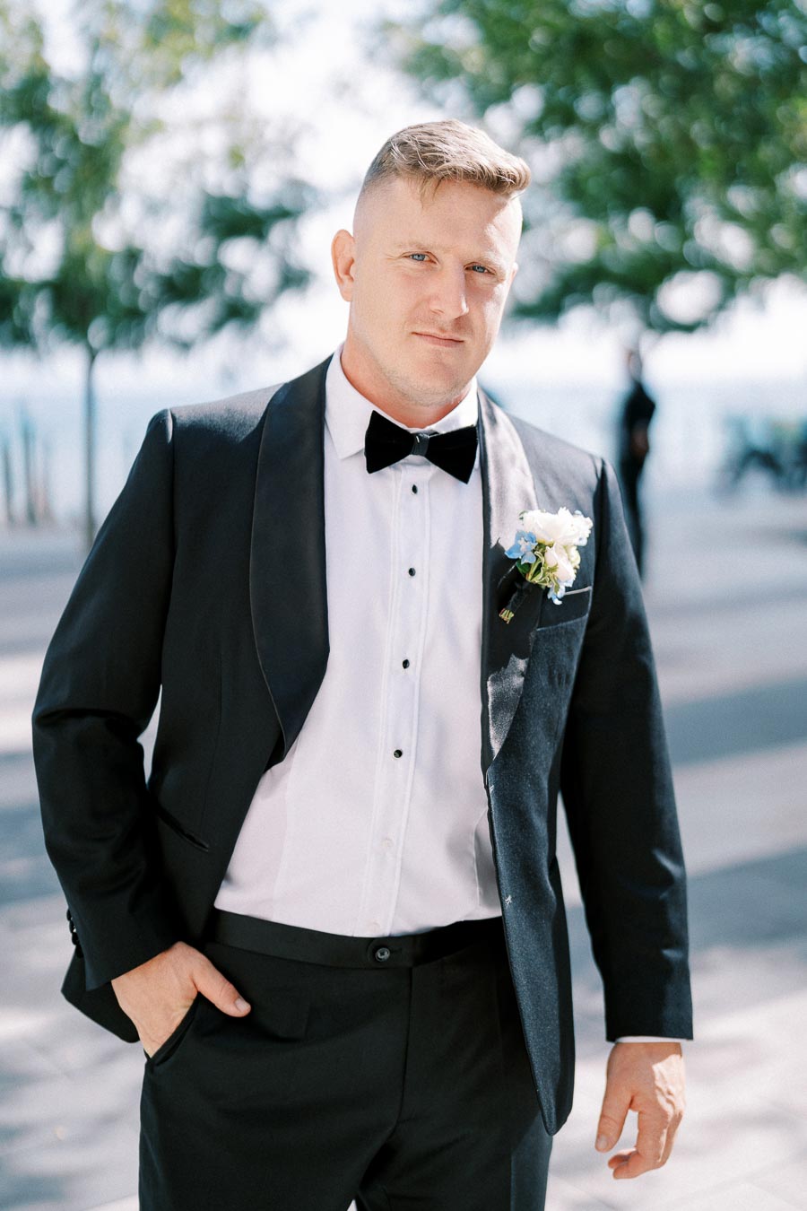 A man in a black tuxedo with a white dress shirt and bow tie, wearing a boutonniere, standing outdoors on a sunny day with trees in the background.