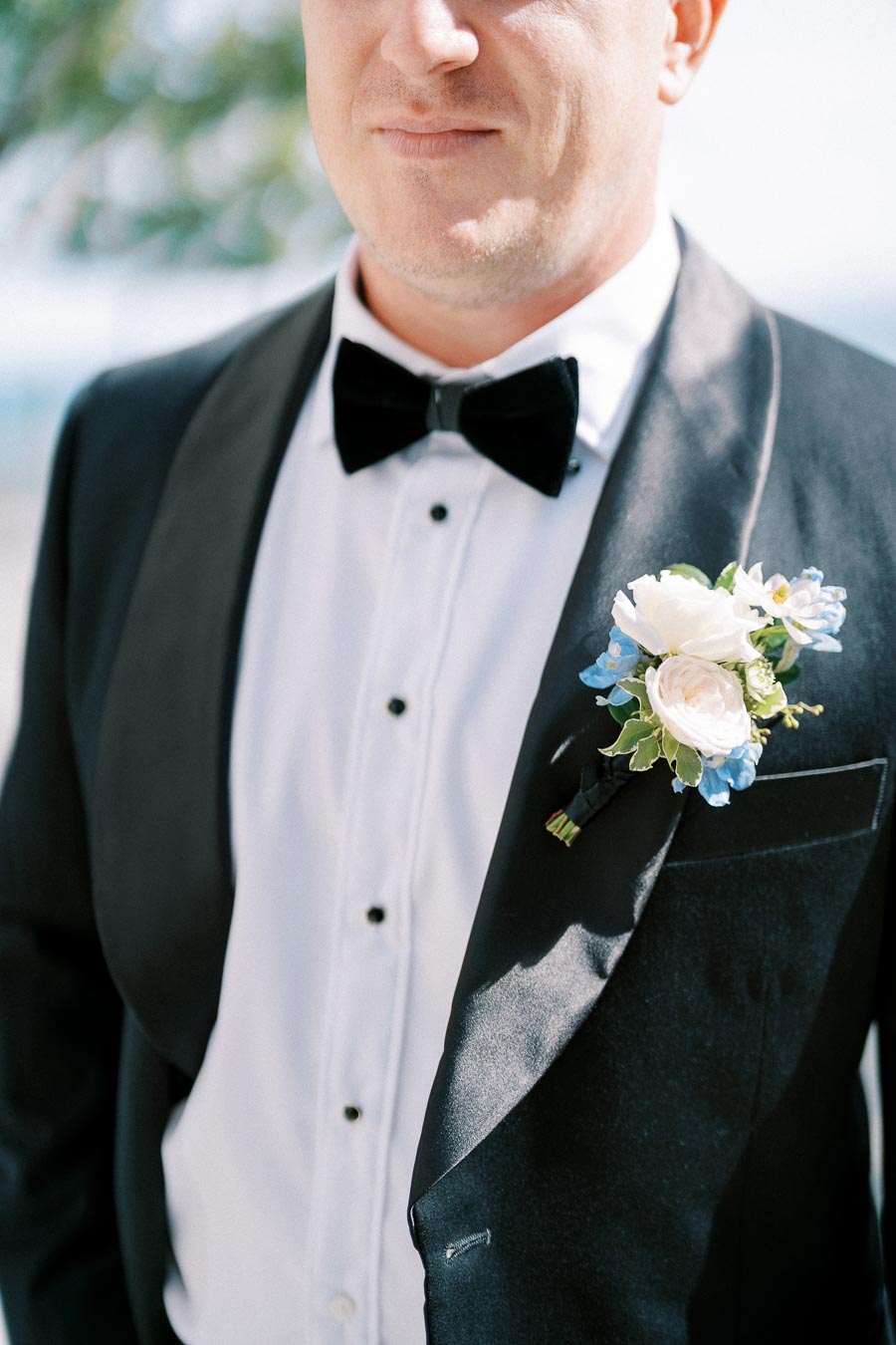 A groom in a black tuxedo and bow tie with a white dress shirt, wearing a boutonniere featuring white and blue flowers, standing outdoors at a wedding.
