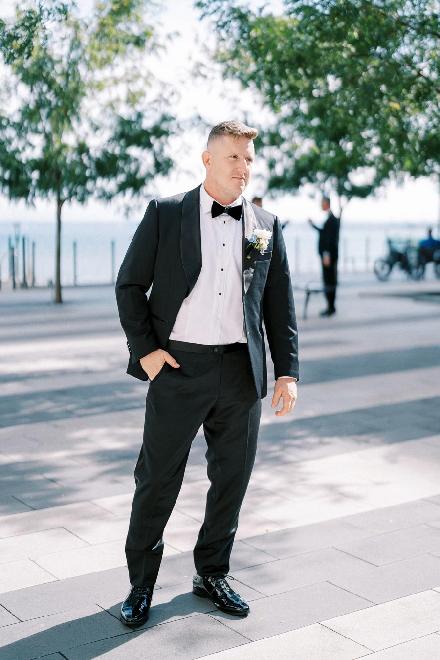 A man in a formal black tuxedo with a bow tie and corsage, standing outdoors on a sunny day with trees and a waterfront in the background.