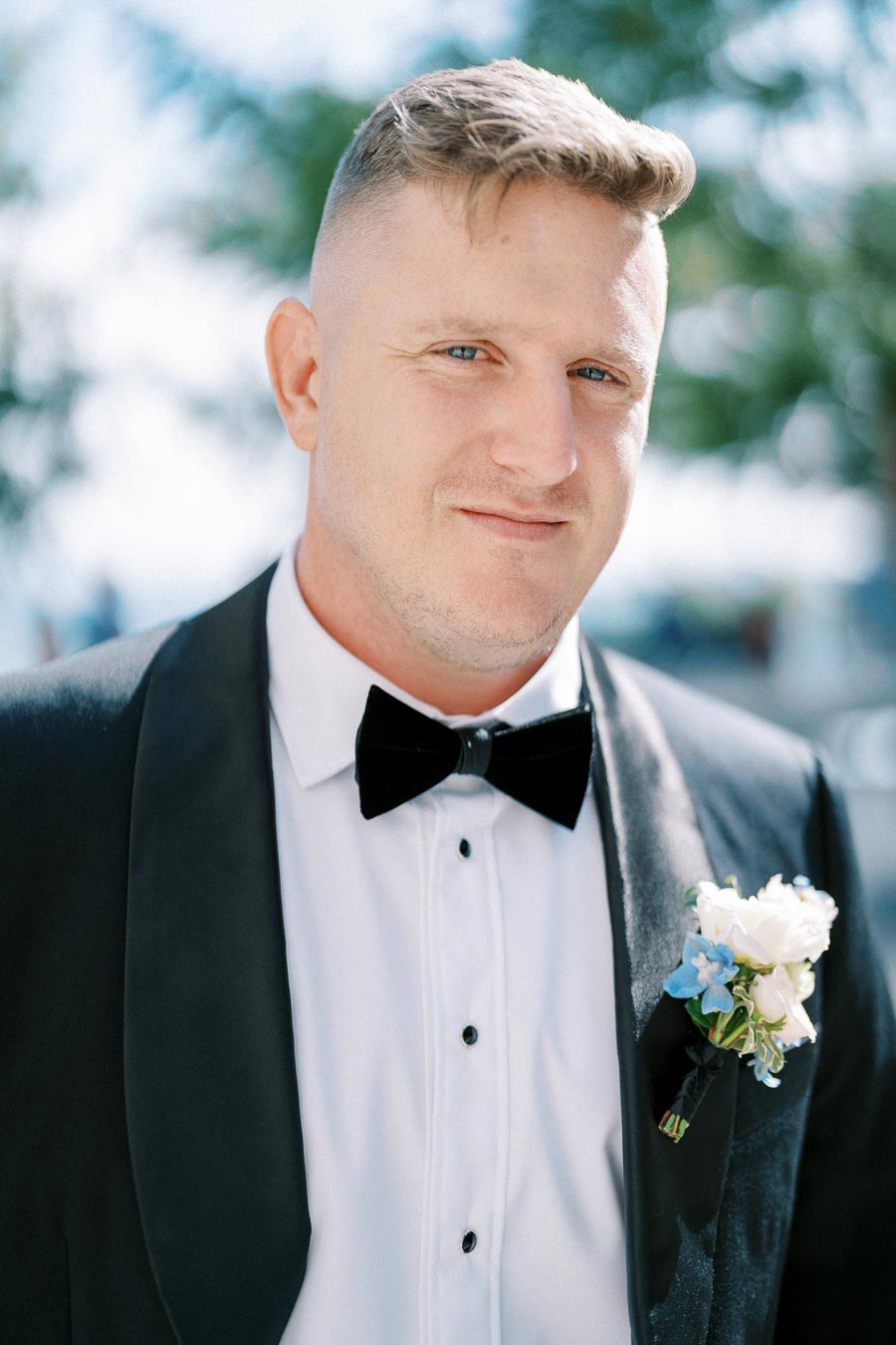 A man in a tuxedo and bow tie, adorned with a white boutonniere, standing outdoors with a blurred natural background.