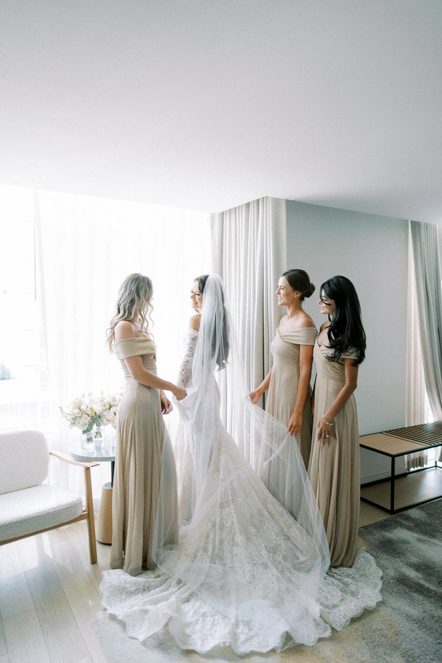 Bridal party getting ready in a bright room, featuring the bride in a lace wedding gown with a long veil, surrounded by three bridesmaids in matching beige dresses, standing by a window with a floral arrangement.