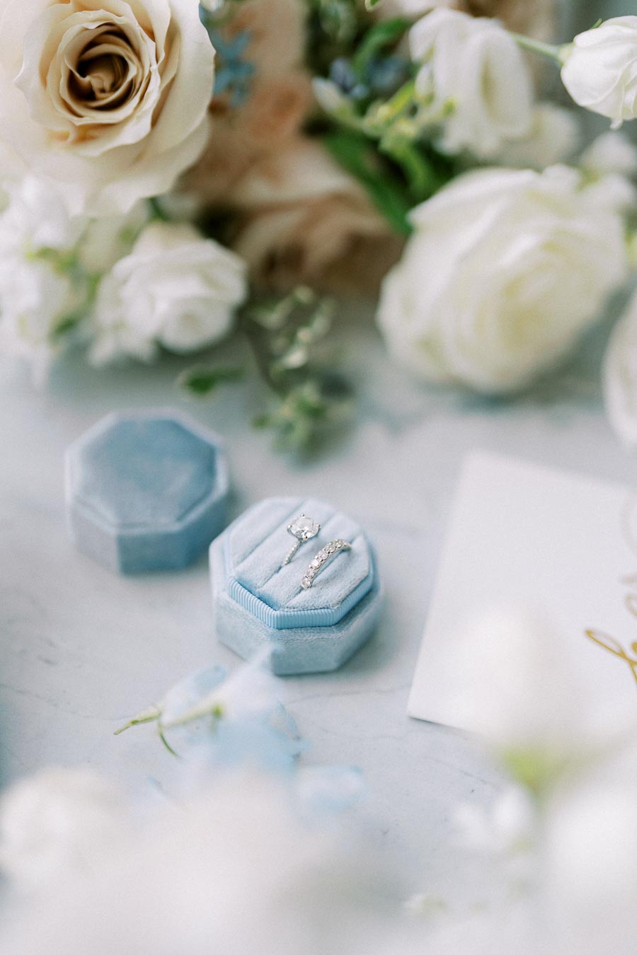 Delicate bridal jewelry set featuring an engagement ring and wedding band in a light blue velvet box, surrounded by elegant white roses and greenery on a marble background.