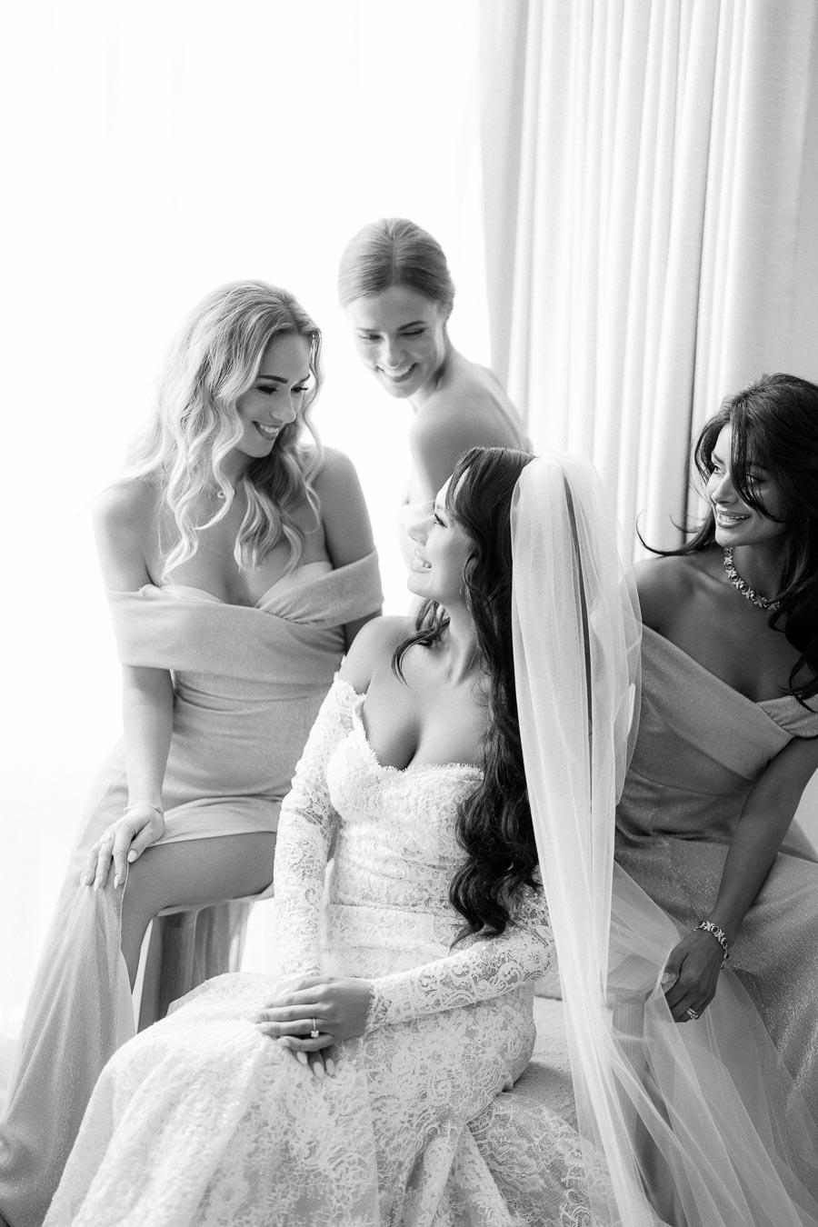 Black and white photo of a bride in a lace wedding dress and veil, surrounded by three bridesmaids in off-shoulder gowns, sharing a joyful moment near a sunlit window.