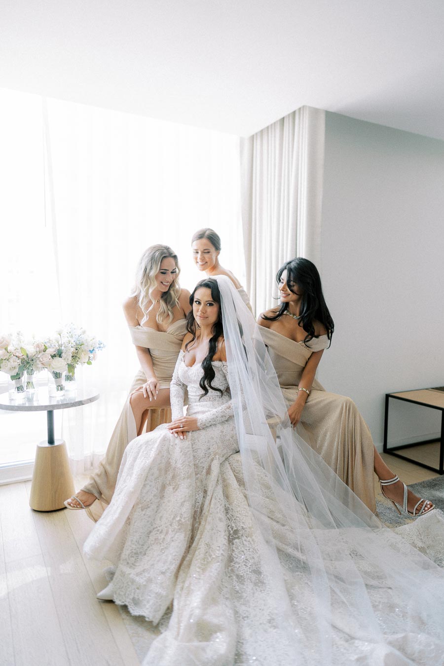 Bridal party in elegant gowns with smiling bridesmaids and bride in lace wedding dress and veil, posing indoors with floral decor.
