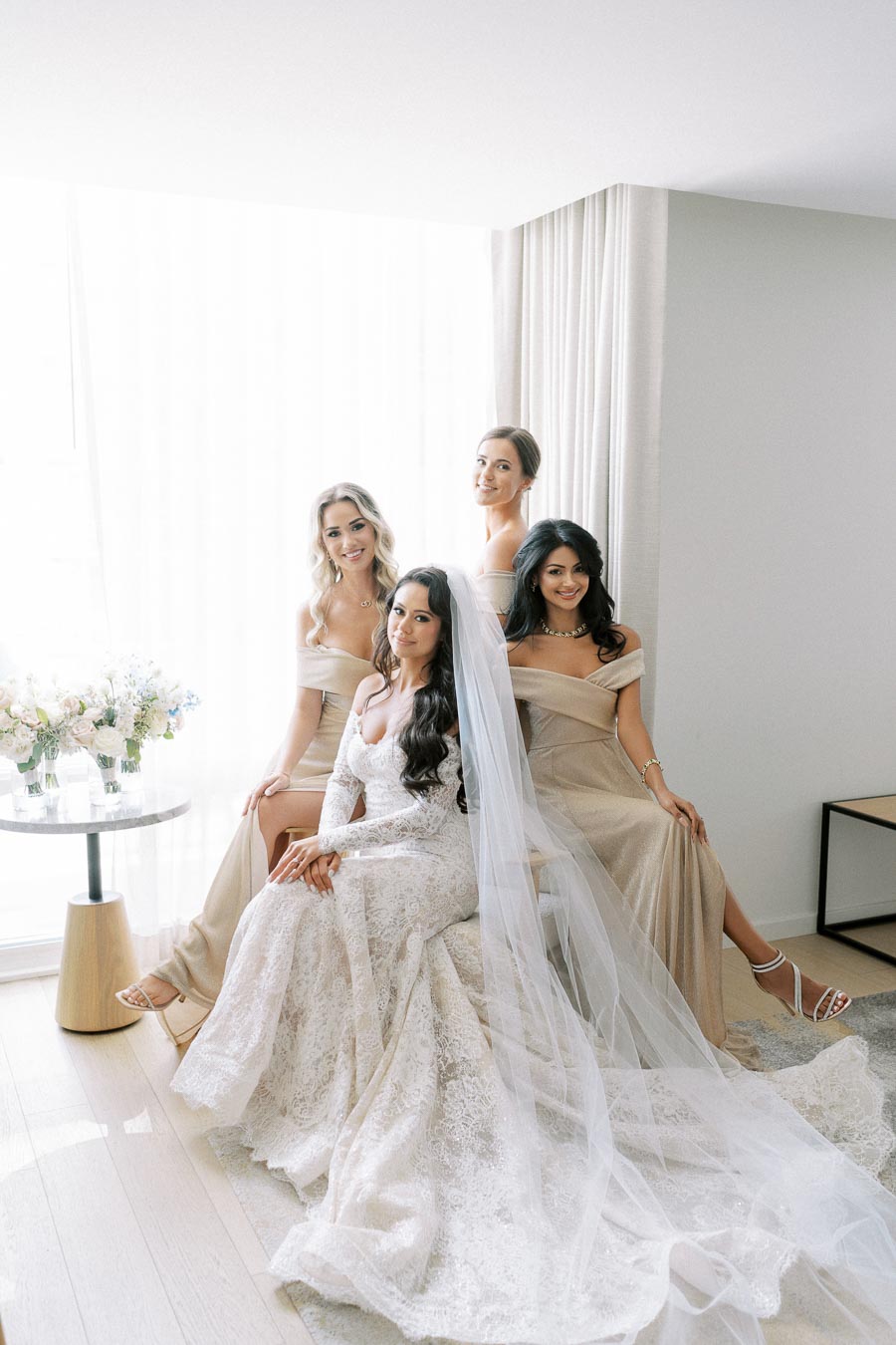 A bride in a lace wedding dress and veil sits with three bridesmaids in gold dresses, posing elegantly in a bright room.
