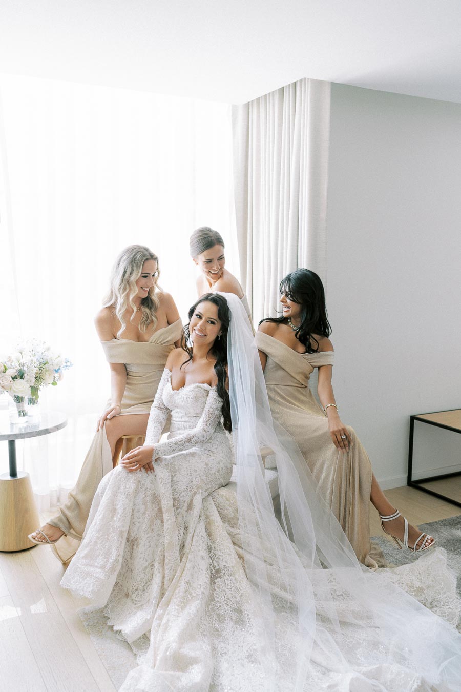 Bride in elegant lace wedding gown sits with bridesmaids in matching champagne dresses, smiling and posing indoors near a window with soft natural light.