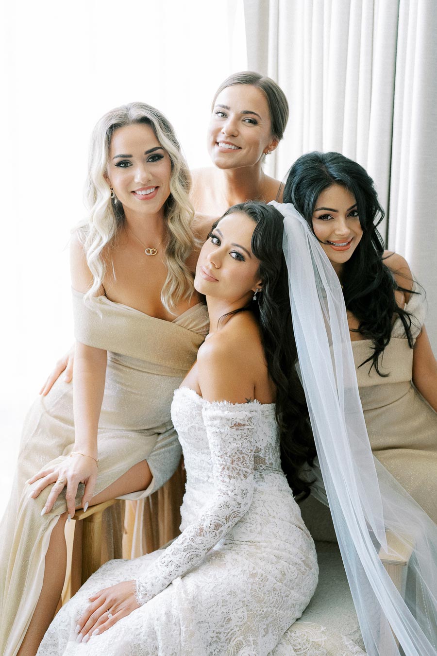Bridal party posing with bride in a white lace wedding dress and veil, accompanied by three bridesmaids in elegant beige dresses, smiling indoors against a soft, neutral backdrop.