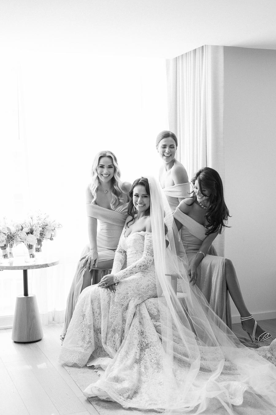 Black and white photo of a smiling bride in an elegant lace wedding dress sitting with her bridesmaids, all dressed in coordinating gowns, in a softly lit room.