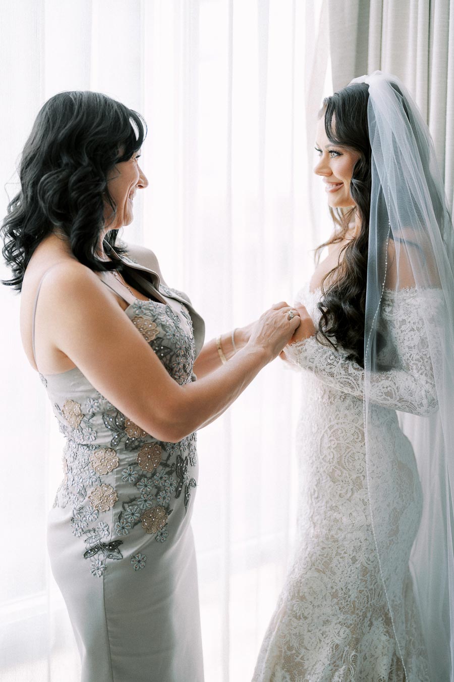 Mother and bride sharing a joyful moment before the wedding ceremony, with the bride in an elegant lace gown and veil, standing by a softly lit window.