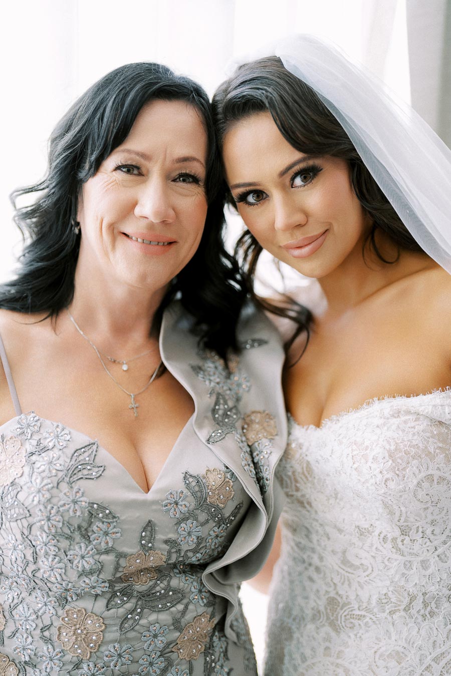 A bride in an elegant lace wedding gown poses with a woman in a floral embroidered dress. Both are smiling and appear close, likely on a wedding day.