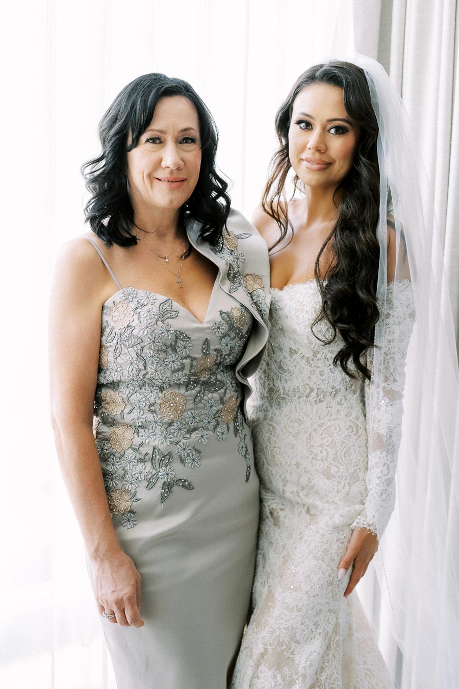 Mother and bride posing together near a window, wearing elegant dresses; the mother in a floral applique gown and the bride in a lace wedding dress with a veil.
